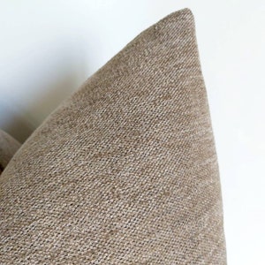 May include: Close-up of a beige throw pillow with a textured fabric. The pillow is made of a soft, woven material.