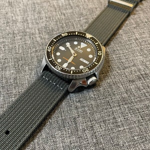 K&ouml;nnte beinhalten: Eine schwarze und silberne Seiko Automatikuhr mit einem schwarzen Nylonarmband. Das Zifferblatt der Uhr hat einen schwarzen Hintergrund mit wei&szlig;en Markierungen und ein Datumsfenster. Die Uhr hat eine drehbare L&uuml;nette mit wei&szlig;en Markierungen.