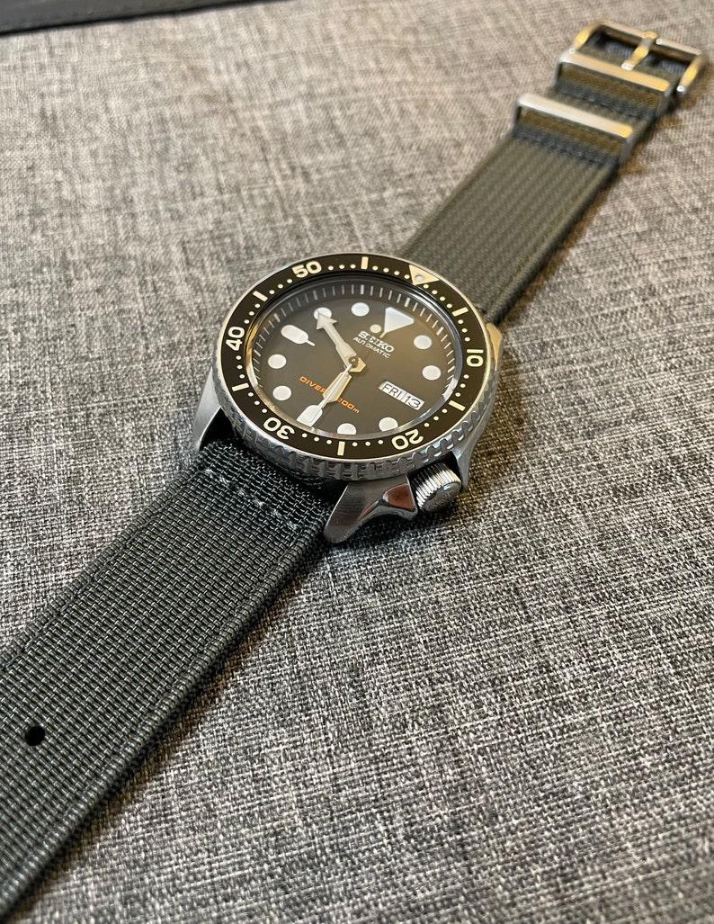 K&ouml;nnte beinhalten: Eine schwarze und silberne Seiko Automatikuhr mit einem schwarzen Nylonarmband. Das Zifferblatt der Uhr hat einen schwarzen Hintergrund mit wei&szlig;en Markierungen und ein Datumsfenster. Die Uhr hat eine drehbare L&uuml;nette mit wei&szlig;en Markierungen.