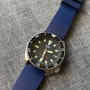 Puede incluir: Un reloj autom&aacute;tico Seiko con esfera negra y correa de silicona azul. El reloj tiene un bisel de acero inoxidable con marcas blancas y una pantalla de fecha a las 3 en punto. El reloj es resistente al agua.