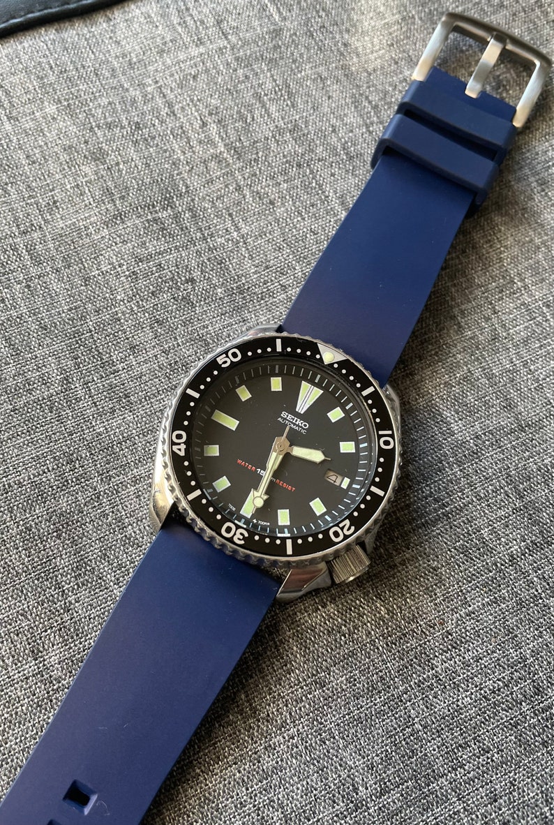 Puede incluir: Un reloj autom&aacute;tico Seiko con esfera negra y correa de silicona azul. El reloj tiene un bisel de acero inoxidable con marcas blancas y una pantalla de fecha a las 3 en punto. El reloj es resistente al agua.