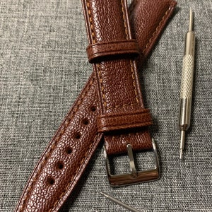 May include: A brown leather watch strap with a silver buckle and two spring bars. The strap has a textured surface and is stitched with a contrasting thread.