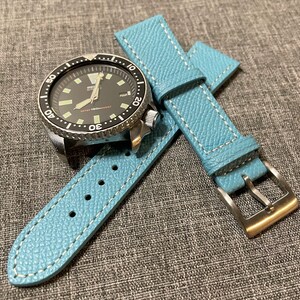 Puede incluir: Una correa de reloj de cuero azul claro con costuras blancas y una hebilla plateada. La correa se muestra con un reloj plateado con una esfera negra y un bisel giratorio.