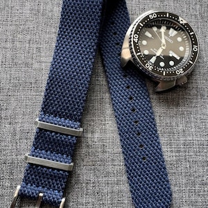 Könnte beinhalten: Ein dunkelblaues Nylon-Armband mit silberner Hardware und einer Schließe. Das Armband ist strukturiert und hat eine gewebte Optik.