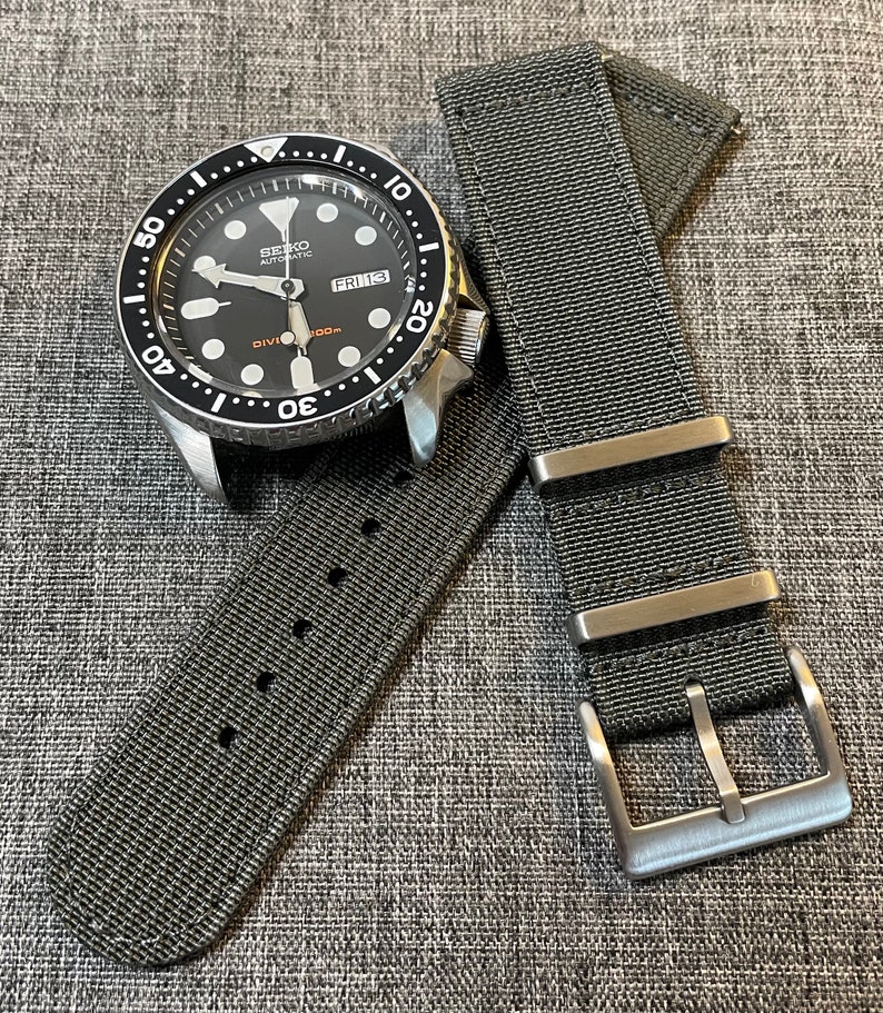 K&ouml;nnte beinhalten: Eine schwarze und silberne Seiko Automatik-Taucheruhr mit einem schwarzen Nylon-Armband. Das Zifferblatt der Uhr hat ein Datumsfenster und der Text "FRI 13" ist sichtbar. Das Armband der Uhr hat eine silberne Schlie&szlig;e.