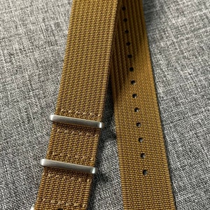 K&ouml;nnte beinhalten: Ein braunes Nylon-Uhrenarmband mit silberner Hardware und einer Schlie&szlig;e. Das Armband hat ein strukturiertes, gewebtes Muster.