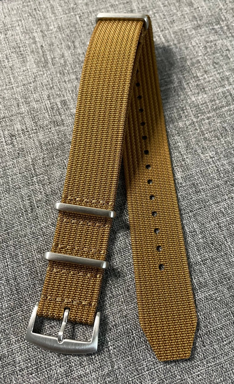 K&ouml;nnte beinhalten: Ein braunes Nylon-Uhrenarmband mit silberner Hardware und einer Schlie&szlig;e. Das Armband hat ein strukturiertes, gewebtes Muster.