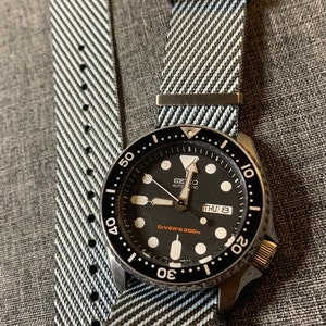 May include: A black and silver Seiko automatic watch with a black face and white markings. The watch has a black and white striped nylon watch band. The watch face says "Diver's 200m" and "Thu 8".