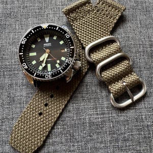 May include: A stainless steel Seiko automatic watch with a black dial and luminous markers, paired with an olive green canvas watch strap. The watch features a rotating bezel and a date display at the 3 o'clock position.