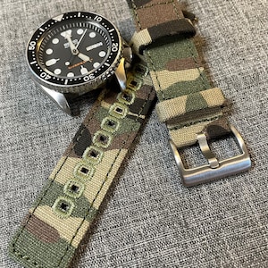 May include: A green, brown, and tan camouflage patterned watch strap with a silver buckle. The strap has a row of small, white squares sewn along the center.