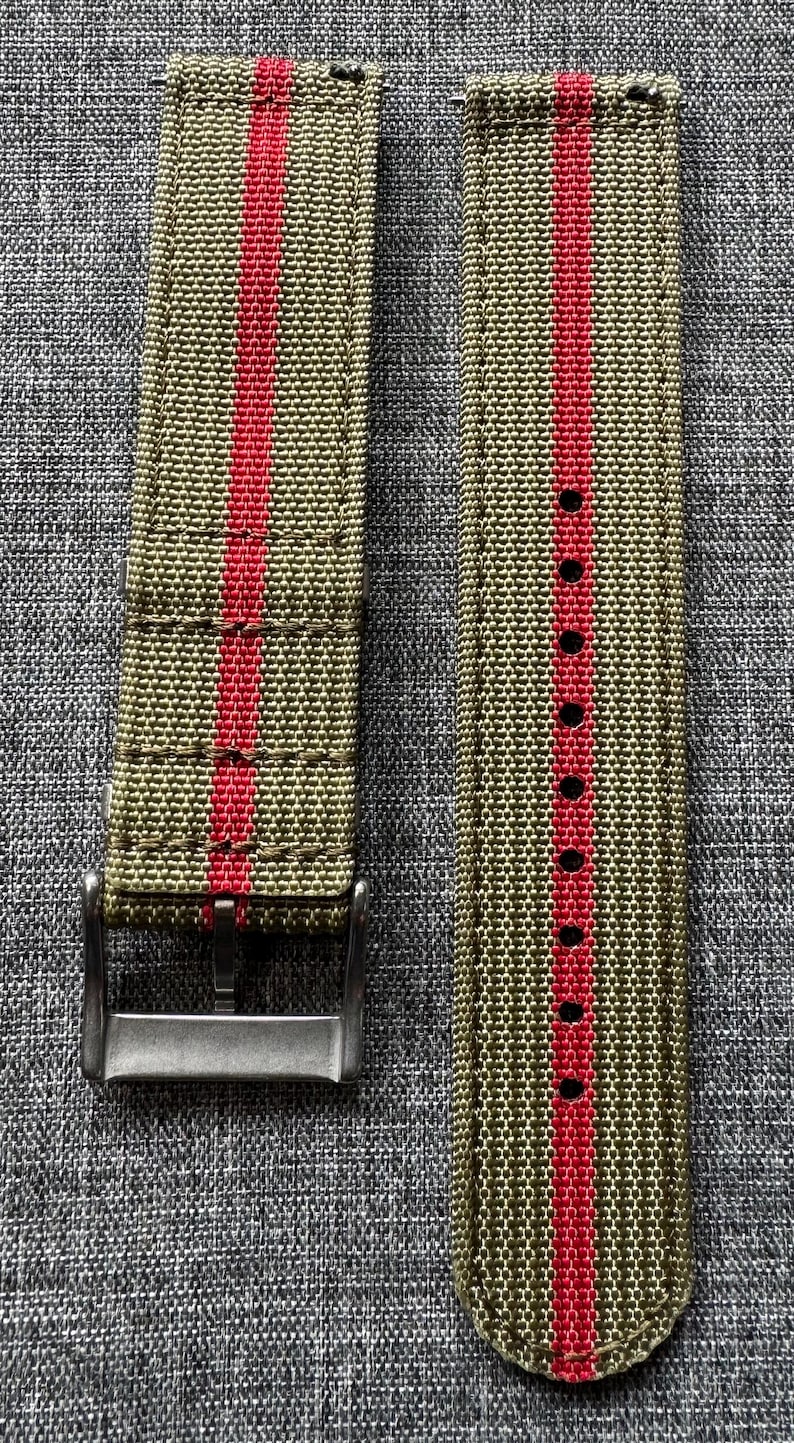 May include: A pair of watch straps with a green and red striped pattern. The straps are made of a woven fabric and have a metal buckle.
