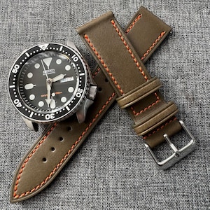 May include: A black-faced Seiko automatic dive watch with a silver bezel and a brown leather watch strap with orange stitching. The watch shows the day and date.
