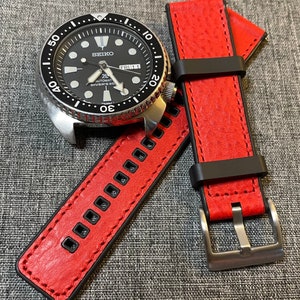 Op de afbeelding: Een rood leren horlogebandje met zwarte stiksels en een zilveren gesp. Het bandje heeft een gestructureerd oppervlak en is ontworpen voor een polshorloge.