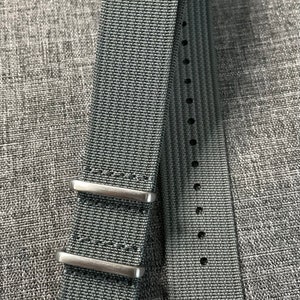 May include: A gray nylon watch strap with silver buckle and keeper loops. The strap has a textured woven pattern.