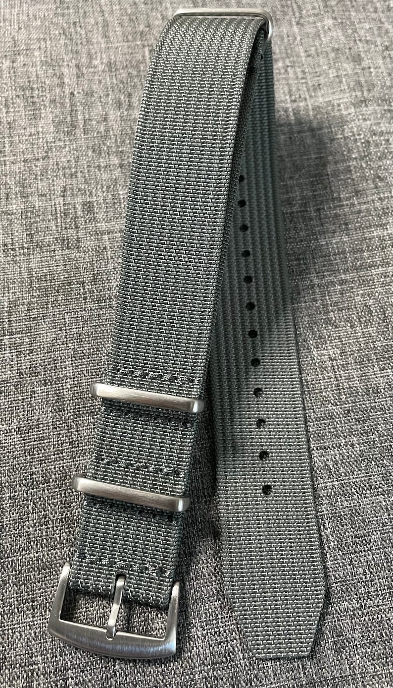 May include: A gray nylon watch strap with silver buckle and keeper loops. The strap has a textured woven pattern.