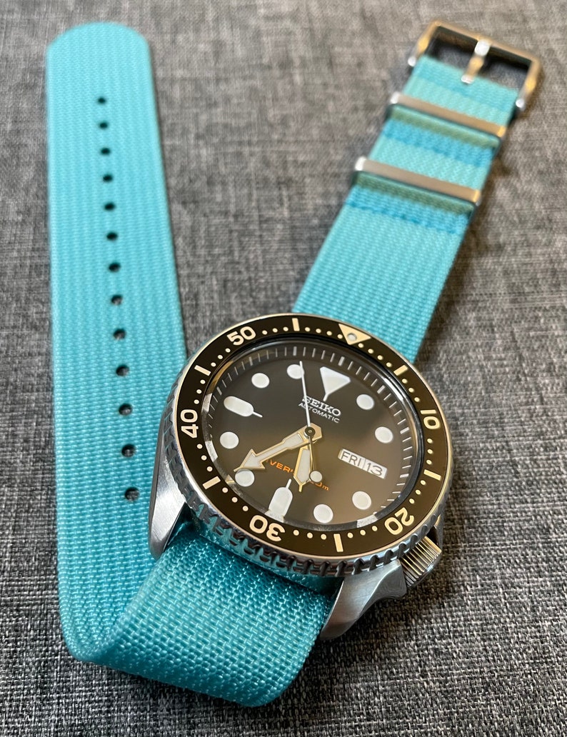 May include: A black and silver wristwatch with a blue nylon watch strap. The watch face has white markings and a date window that reads "FRI 13".