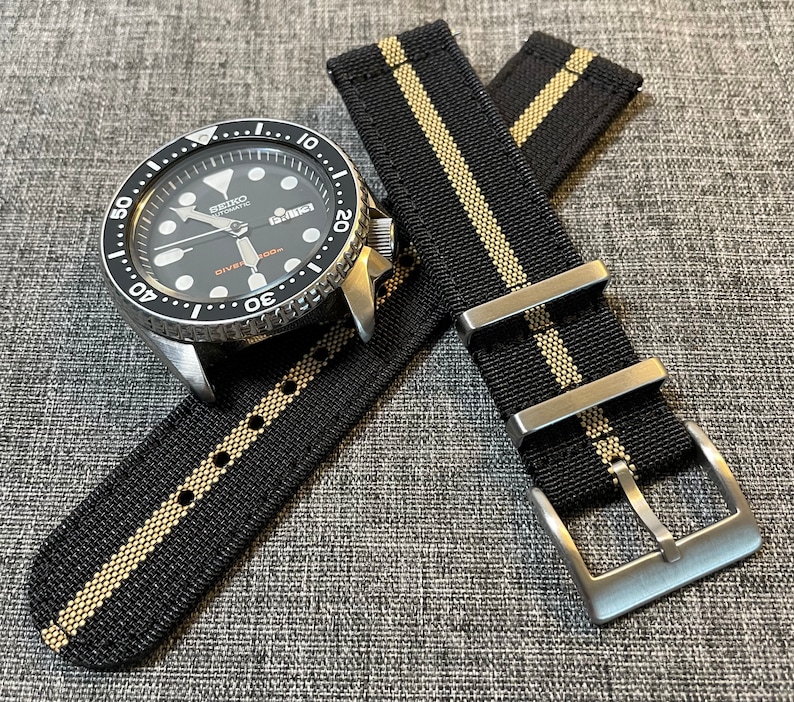 May include: A black and tan striped watch strap with a silver buckle. The strap is made of nylon and has a textured finish. The strap is shown on a black and silver watch with a black face and white markings.