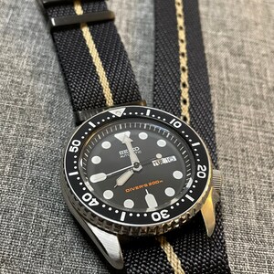 May include: A black and silver wristwatch with a black face and white markings. The watch has a black and white striped nylon strap. The watch face reads "SEIKO AUTOMATIC" and "DIVER'S 200m". The date window shows "TUE 16".