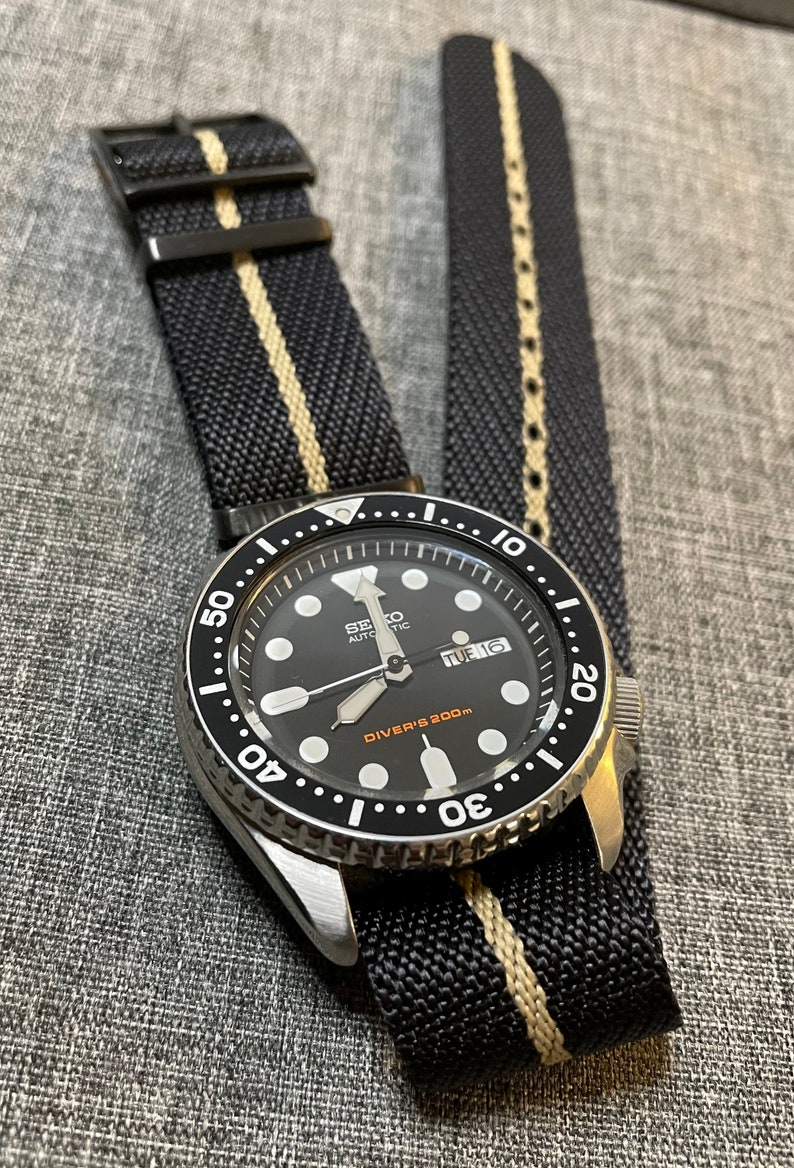 May include: A black and silver wristwatch with a black face and white markings. The watch has a black and white striped nylon strap. The watch face reads "SEIKO AUTOMATIC" and "DIVER'S 200m". The date window shows "TUE 16".
