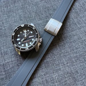 Könnte beinhalten: Eine schwarze und silberne Armbanduhr mit schwarzem Zifferblatt und grauem Silikonarmband. Die Uhr hat eine drehbare Lünette und eine Datumsanzeige. Das Armband ist mit einer silbernen Schließe verbunden.