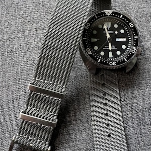 May include: A gray nylon watch strap with a silver buckle. The strap is laying on a gray fabric surface next to a black and silver Seiko watch with a black face and white markings. The watch face has the text "Automatic Diver's 200m" and the date window says "SAT 7".