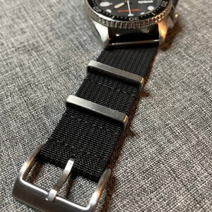 May include: A black nylon watch strap with a silver buckle. The strap is attached to a silver watch with a black face and a silver bezel.