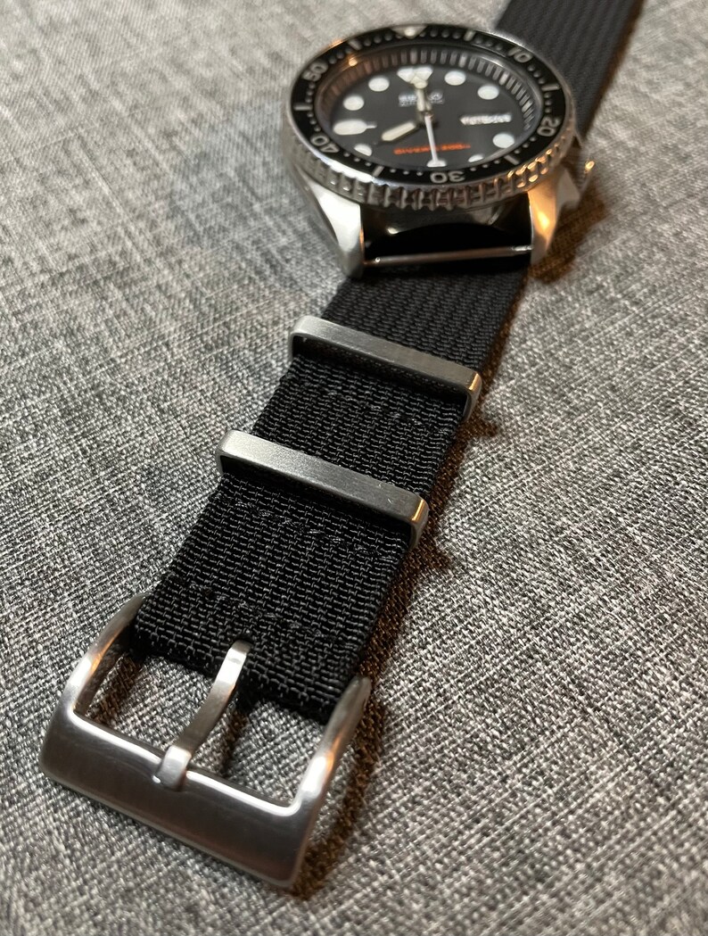 May include: A black nylon watch strap with a silver buckle. The strap is attached to a silver watch with a black face and a silver bezel.