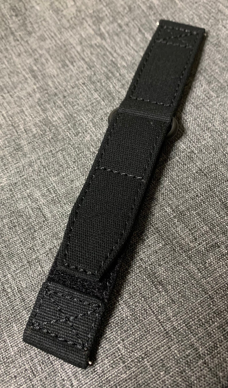 May include: Black watch strap with a metal buckle. The strap is made of a woven fabric.