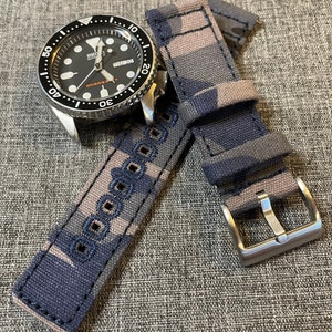 May include: A black and grey camouflage watch strap with a silver buckle. The strap has a black stitching detail and is made of canvas.