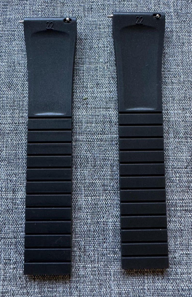 Puede incluir: Dos correas de reloj negras con la marca 22. Las correas est&aacute;n hechas de un material flexible y tienen un dise&ntilde;o segmentado. Se muestran sobre un fondo gris texturizado.