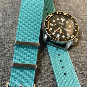 May include: A light blue nylon watch strap with silver hardware and a black watch face with white markings. The watch face reads "SEIKO AUTOMATIC".