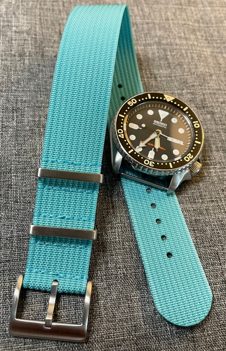May include: A light blue nylon watch strap with silver hardware and a black watch face with white markings. The watch face reads "SEIKO AUTOMATIC".