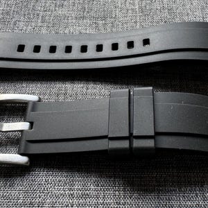 May include: Two-piece black watch band with a stainless steel buckle. The top piece has multiple rectangular holes. The band is made of a flexible material and is designed to attach to a watch case.