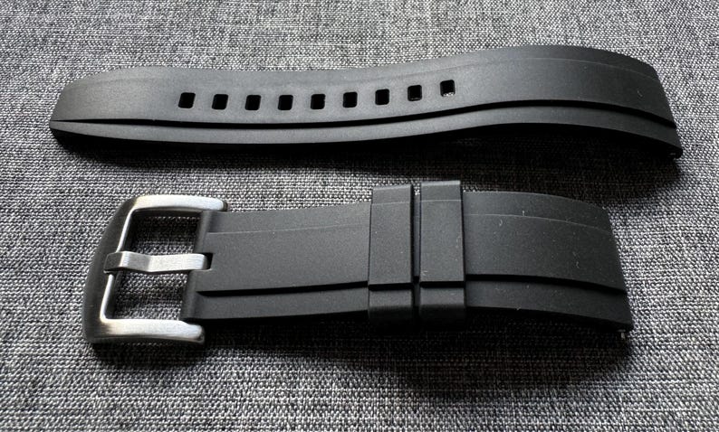 May include: Two-piece black watch band with a stainless steel buckle. The top piece has multiple rectangular holes. The band is made of a flexible material and is designed to attach to a watch case.