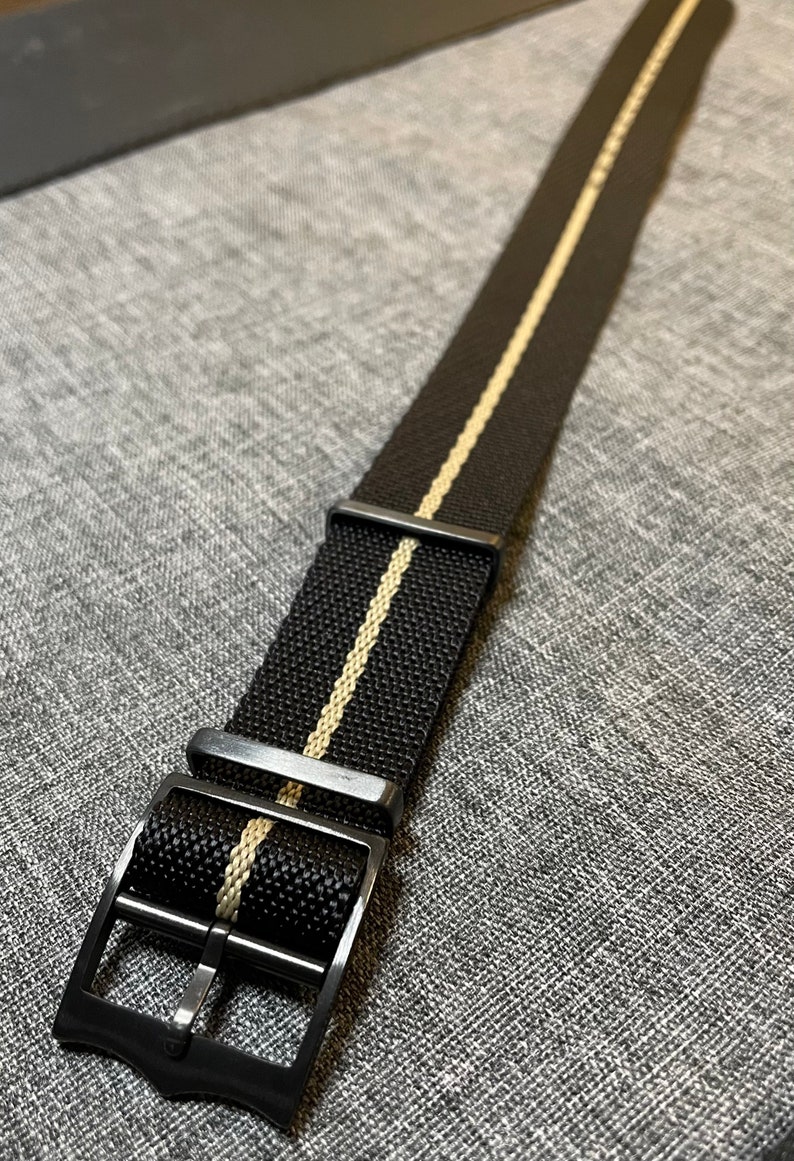 May include: A black watch strap with a white stripe down the center. The strap has a black metal buckle.