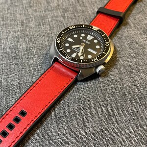 Op de afbeelding: Een zilveren horloge met een zwarte wijzerplaat en een rode leren band. De wijzerplaat heeft witte markeringen en een datumscherm. De band heeft zwarte stiksels.