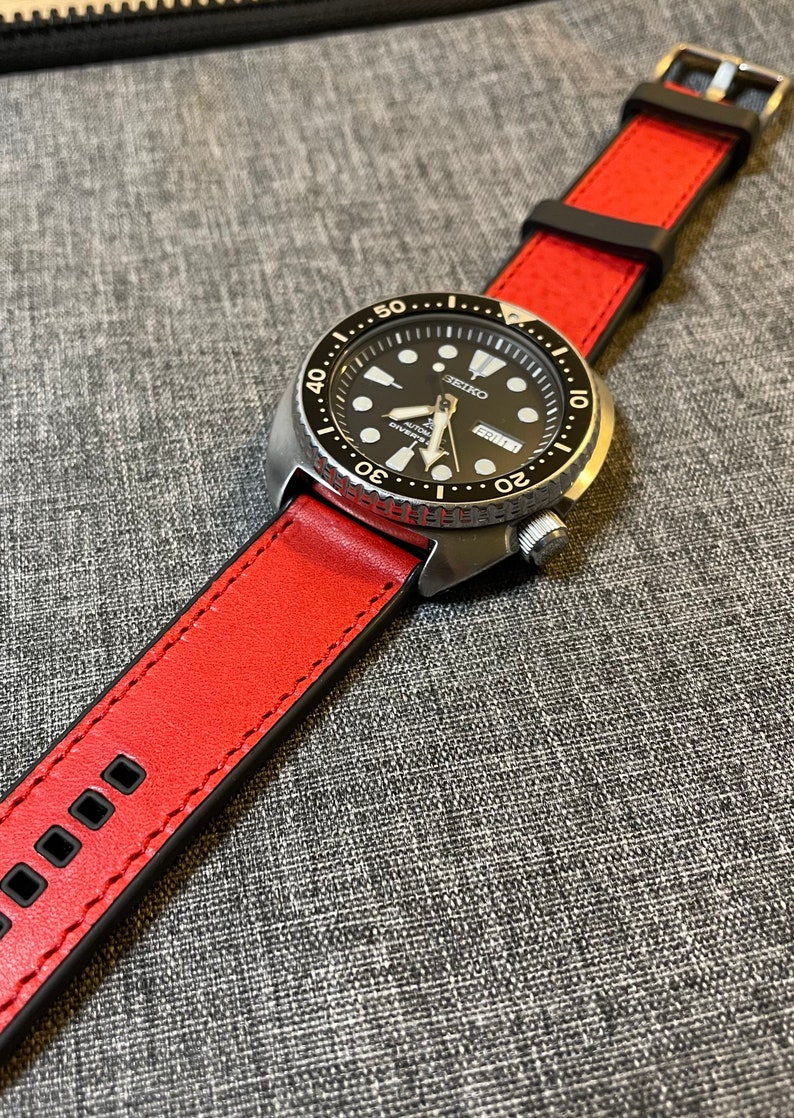 Op de afbeelding: Een zilveren horloge met een zwarte wijzerplaat en een rode leren band. De wijzerplaat heeft witte markeringen en een datumscherm. De band heeft zwarte stiksels.