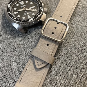 Puede incluir: Una correa de reloj de cuero gris con una hebilla plateada. La correa tiene una superficie texturizada y está cosida con hilo blanco.