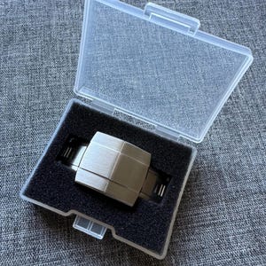 Puede incluir: Un reloj plateado con un dise&ntilde;o geom&eacute;trico se muestra dentro de una caja de pl&aacute;stico transparente con un inserto de espuma negra. El reloj tiene una esfera rectangular y un acabado de metal cepillado. La caja est&aacute; abierta.