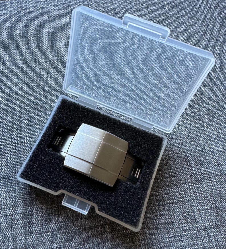 Puede incluir: Un reloj plateado con un dise&ntilde;o geom&eacute;trico se muestra dentro de una caja de pl&aacute;stico transparente con un inserto de espuma negra. El reloj tiene una esfera rectangular y un acabado de metal cepillado. La caja est&aacute; abierta.