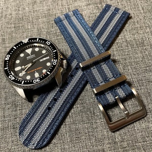 May include: A black and silver Seiko automatic watch with a blue and white striped nylon watch strap. The watch face has a black background with white markings and the text "SEIKO AUTOMATIC DIVER'S 200m". The strap has a silver buckle.