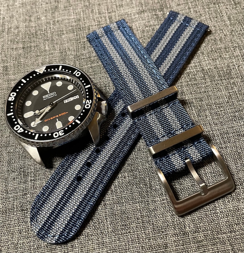 May include: A black and silver Seiko automatic watch with a blue and white striped nylon watch strap. The watch face has a black background with white markings and the text "SEIKO AUTOMATIC DIVER'S 200m". The strap has a silver buckle.