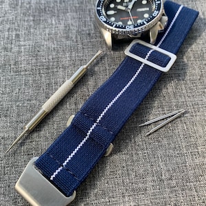 Peut inclure: Un bracelet de montre bleu marine avec des rayures blanches et des accessoires argentés. Le bracelet est posé à plat sur une surface en tissu gris. Il y a deux barrettes à ressort argentées à côté du bracelet.
