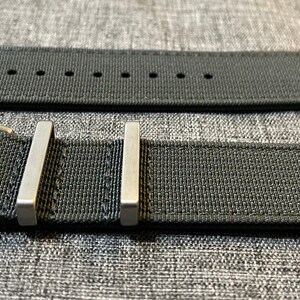 K&ouml;nnte beinhalten: Ein dunkelgraues Nylon-Armband mit einer silbernen Schlie&szlig;e und zwei silbernen Halte&ouml;sen.