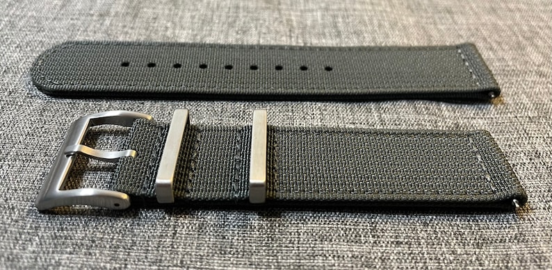 K&ouml;nnte beinhalten: Ein dunkelgraues Nylon-Armband mit einer silbernen Schlie&szlig;e und zwei silbernen Halte&ouml;sen.