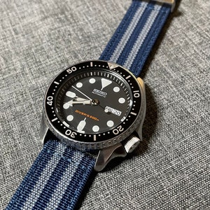 May include: A black Seiko automatic diver's watch with a blue and white striped nylon watch strap. The watch face has a black background with white markings and a date window that reads "FRI 20".