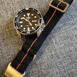 Pode incluir: Um relógio de pulso automático Seiko com mostrador preto, marcadores de hora luminosos e exibição de dia e data. O relógio possui uma luneta rotativa com números brancos e uma pulseira de tecido preto com uma fina faixa central laranja.