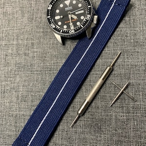May include: A navy blue watch strap with a white stripe down the center. The strap is shown with a watch, a tool for removing watch bands, and two spring bars.