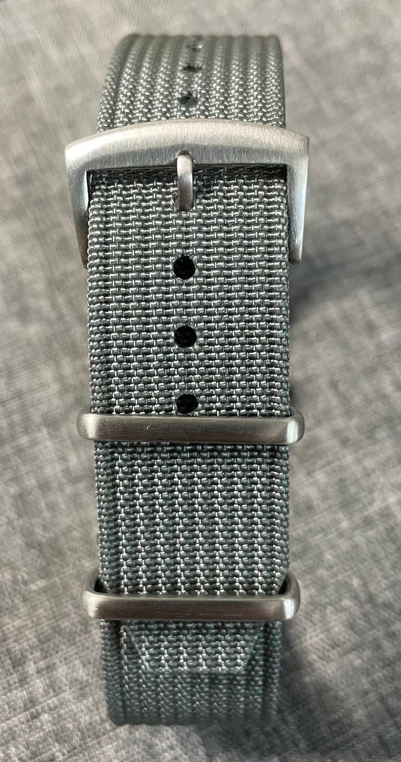 May include: A gray nylon watch strap with a silver buckle and three keeper loops.