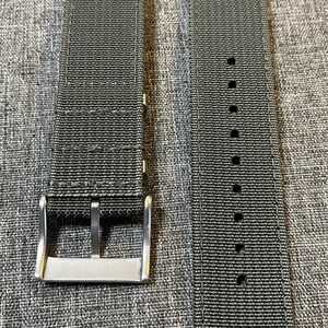 K&ouml;nnte beinhalten: Zwei dunkelgraue Nylon-Uhrenarmb&auml;nder mit silbernen Schlie&szlig;en. Die Armb&auml;nder sind aus gewebtem Material und haben eine strukturierte Oberfl&auml;che.